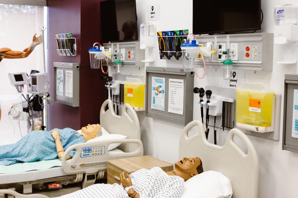 Two medical training mannequins lie in hospital beds in a simulation room, surrounded by monitors, medical equipment, and wall-mounted supplies.