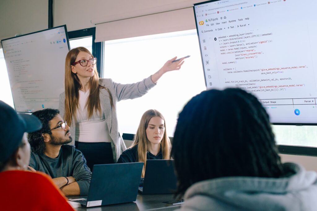 A professor stands and points to computer code on a large screen during a meeting, while three seated individuals look on, each with laptops open.