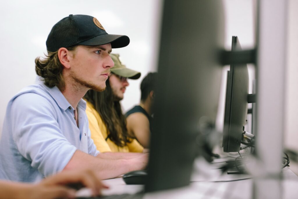 Three people sit side by side at computer monitors, focused on their screens in a classroom or lab setting.