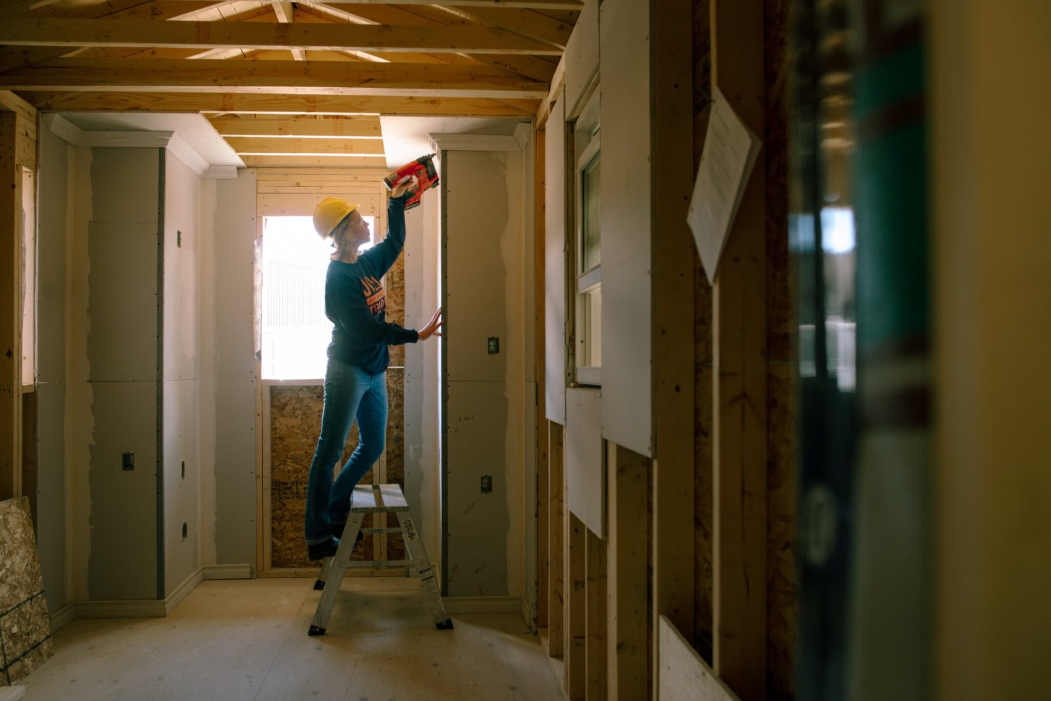 An individual in a hard hat stands on a ladder using a power tool to work on a wooden ceiling in a partially constructed room with exposed framing.