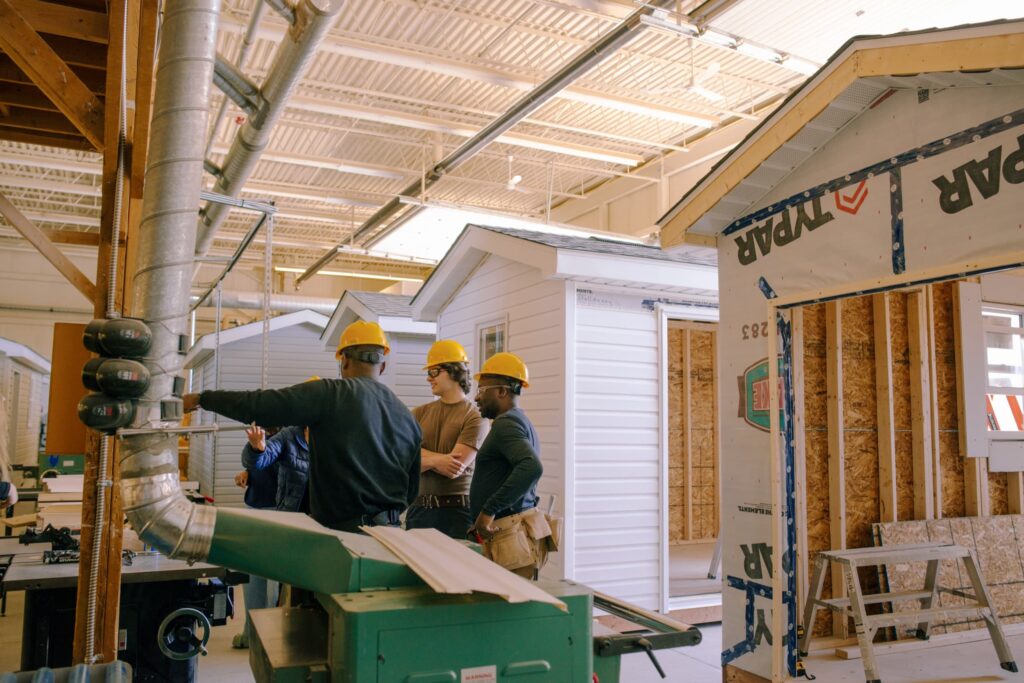 Several people wearing hard hats stand inside a workshop, discussing construction next to small, partially built houses.