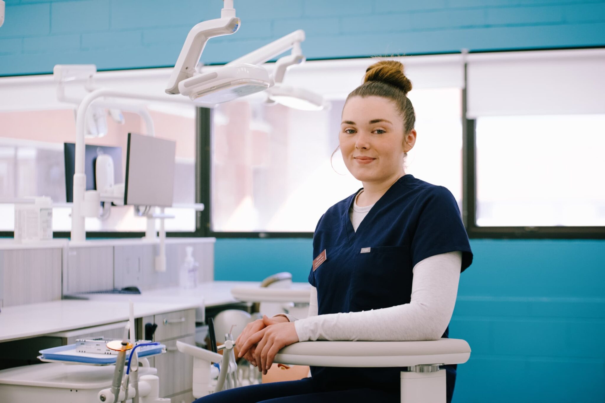A dental hygienist wearing navy scrubs sits on a dental chair in a bright, modern dental clinic with equipment visible in the background.