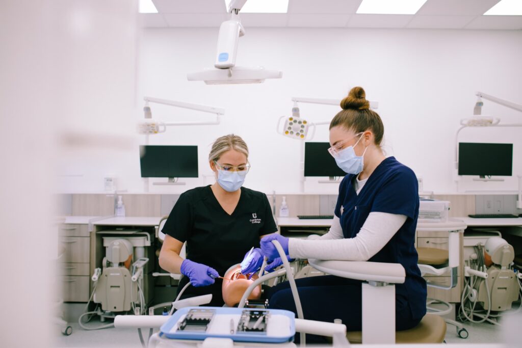Two dental students wearing masks and gloves examine a patient's mouth in a modern dental clinic with computers and equipment visible.