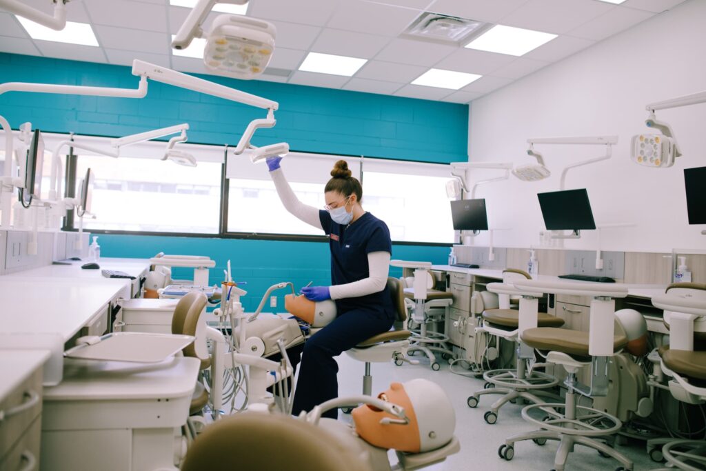 A student wearing scrubs and a mask adjusts equipment in a dental training classroom with empty chairs, dental stations, and computer monitors.