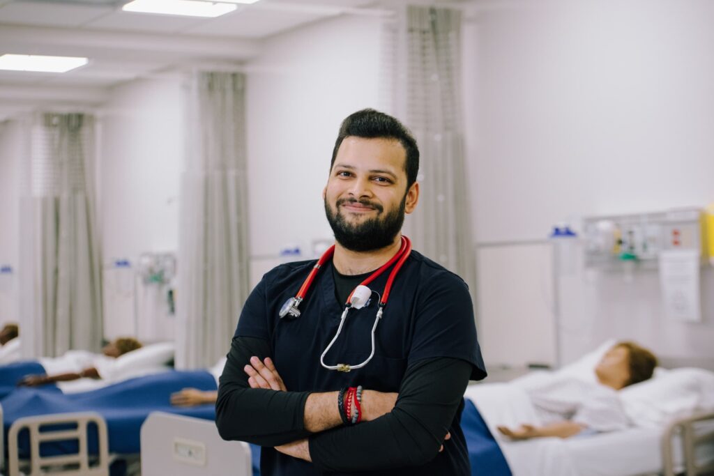 A healthcare professional with a stethoscope stands smiling with arms crossed in a hospital room with patients lying in beds in the background.