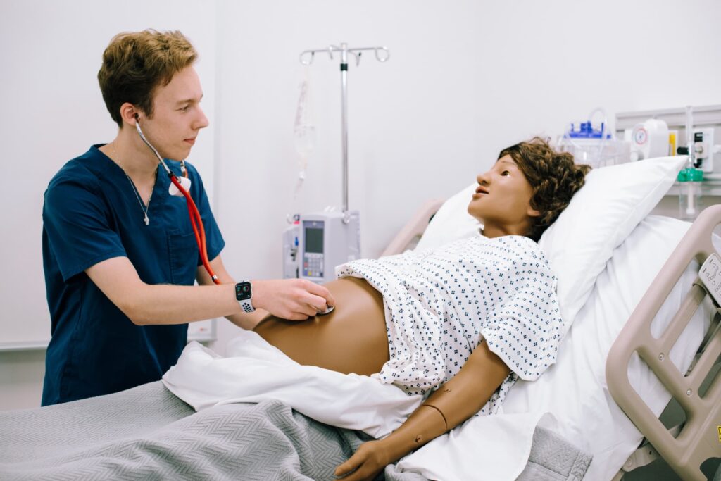 A medical student uses a stethoscope on a patient simulator mannequin lying in a hospital bed during a training exercise.