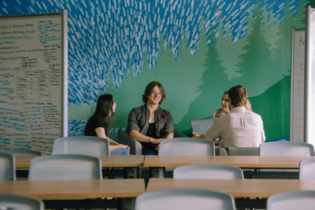Four people sit and talk in a classroom with a forest mural and whiteboards covered in notes, while rows of empty desks are in the foreground.