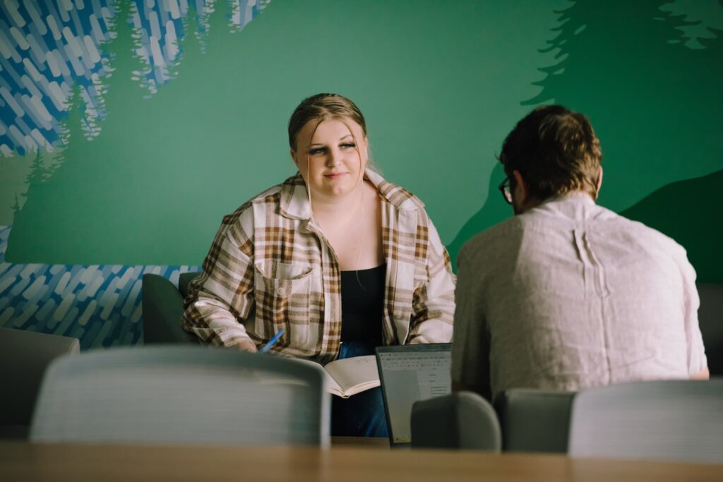 Two people sit across from each other at a table; one holds an open notebook, and the other faces a laptop. A green mural is visible on the wall behind them.