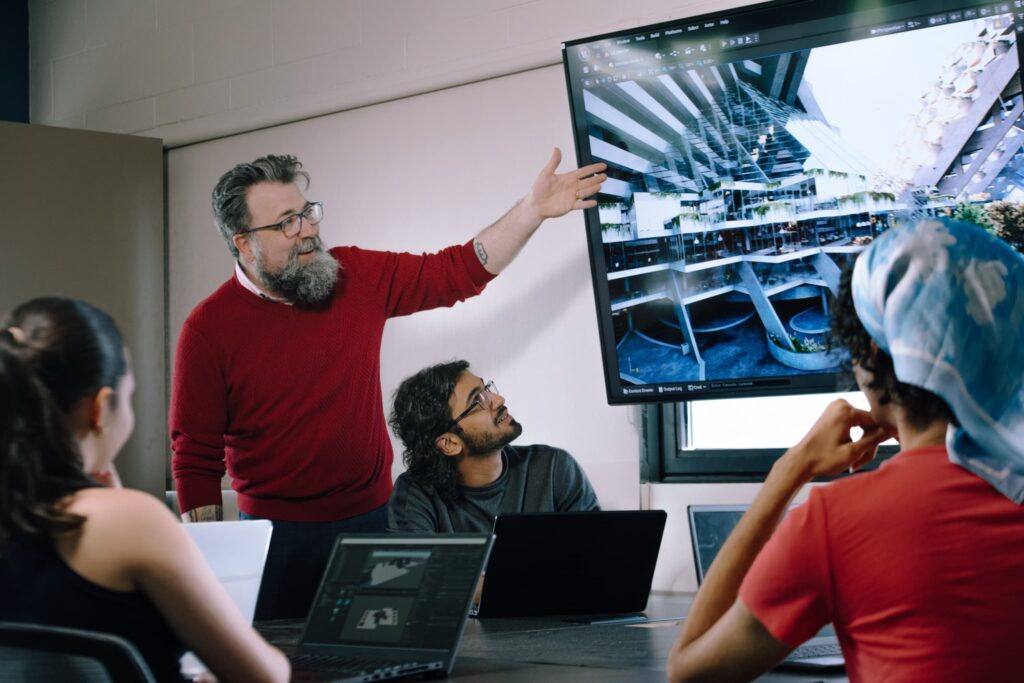 A bearded individual in a red sweater presents architectural designs on a large screen to three colleagues seated at a conference table with laptops.