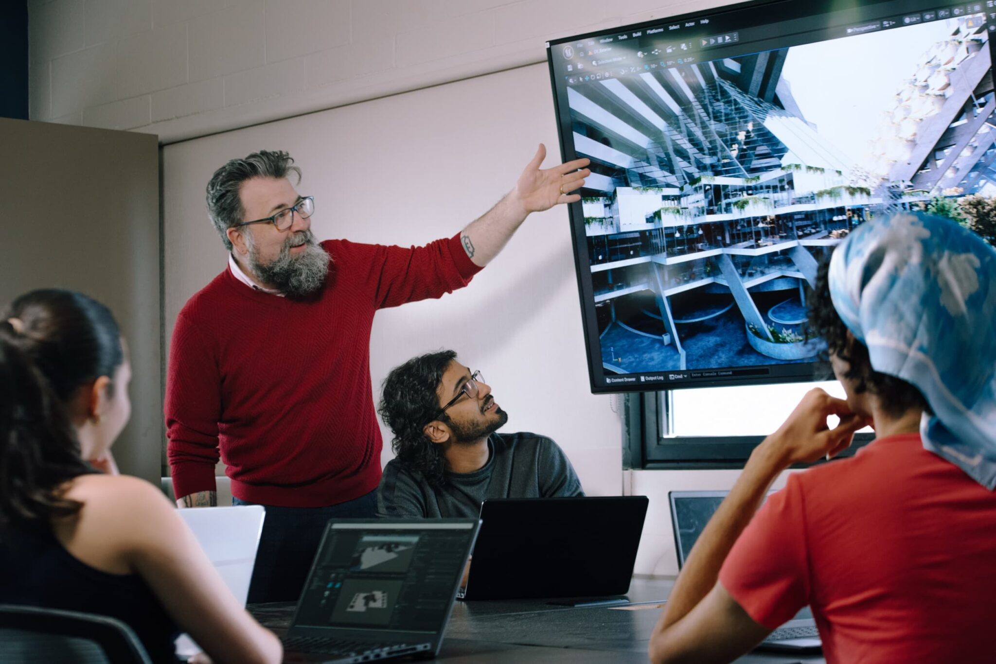 A bearded individual in a red sweater presents architectural designs on a large screen to three colleagues seated at a conference table with laptops.