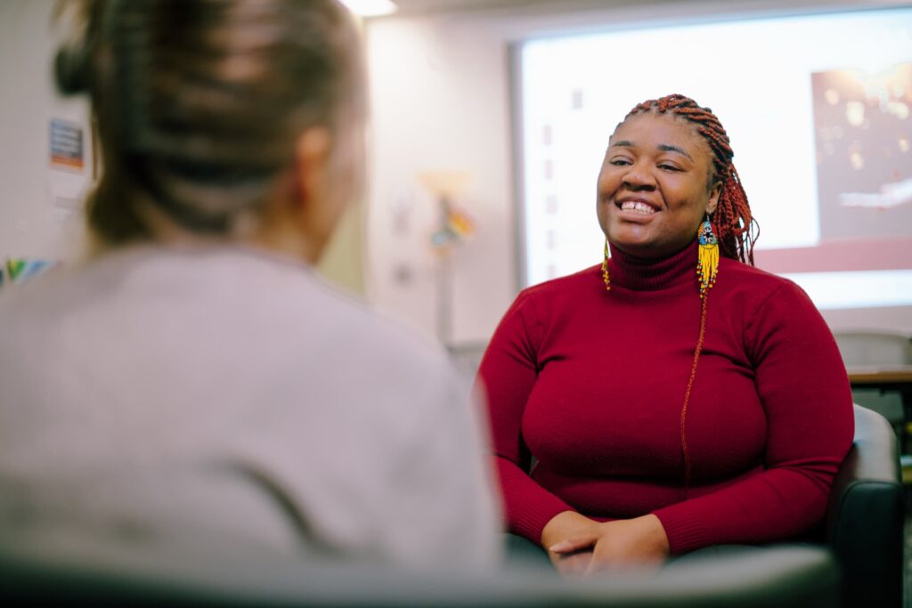 A person in a red sweater sits smiling and talking to another person in an office or classroom setting with a projection screen in the background.