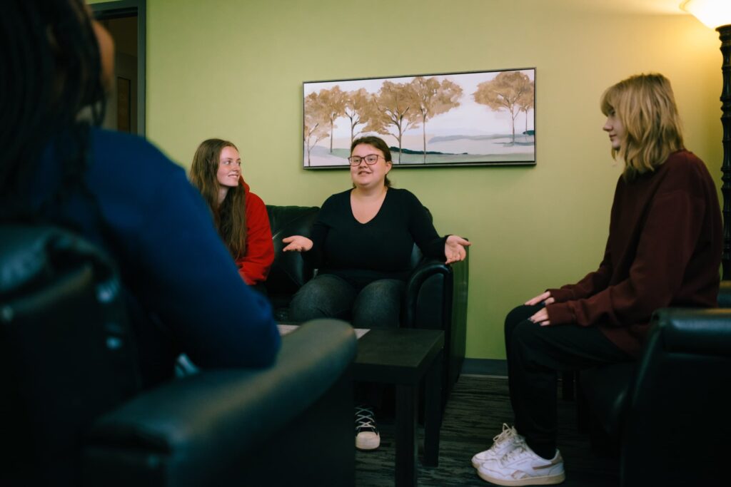 Four people sit in a living room, three facing one person who is speaking with hands outstretched; painting and lamp in the background.