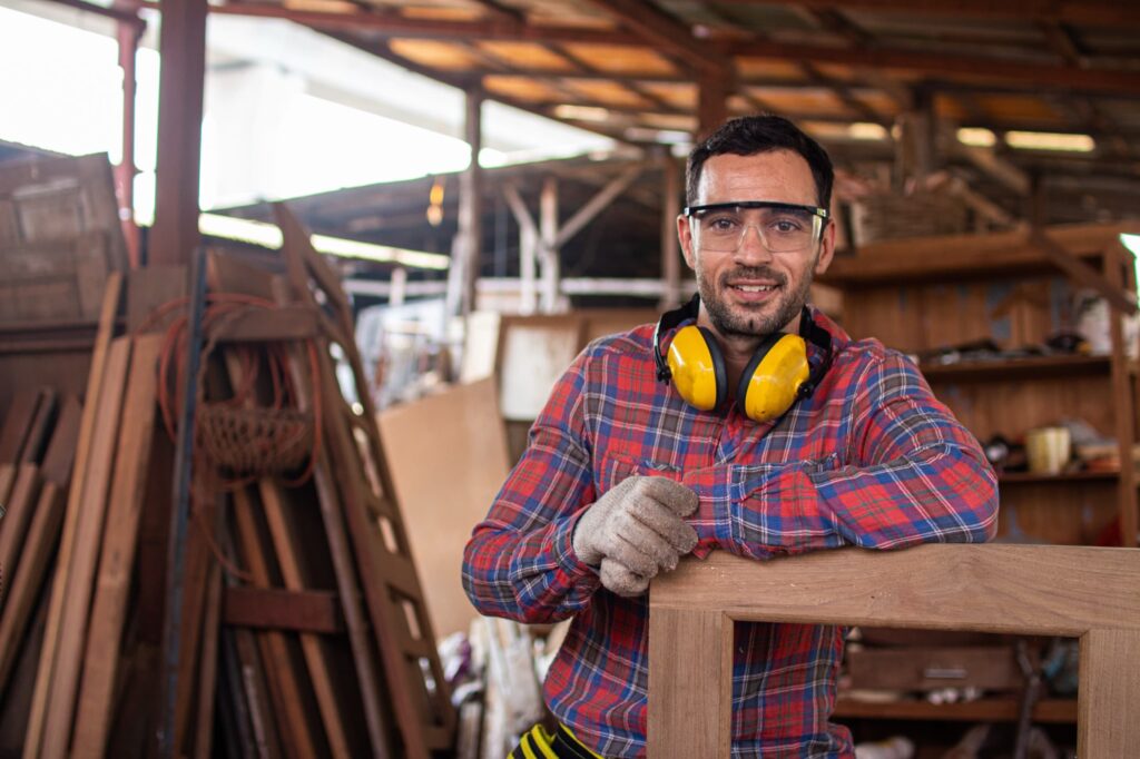 A man wearing safety glasses, gloves, and ear protection stands in a woodworking shop, leaning on a wooden frame. Various pieces of wood and tools are visible in the background.