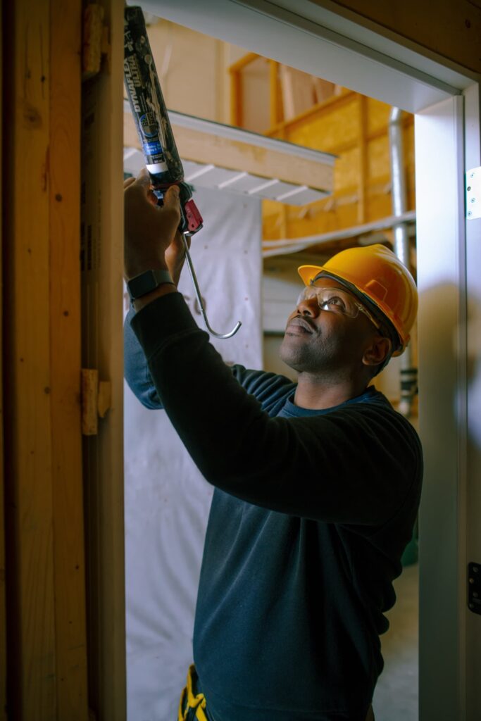 A construction worker in a hard hat and safety glasses uses a caulk gun to apply sealant to a door frame.