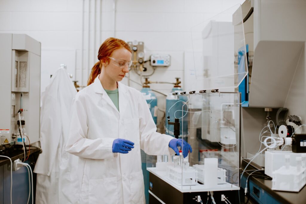 A researcher in a lab coat, safety goggles, and blue gloves works at a laboratory instrument.