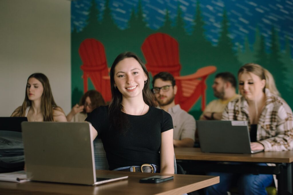 A young woman smiling at a laptop at a desk in a classroom, with other students working at laptops in the background.