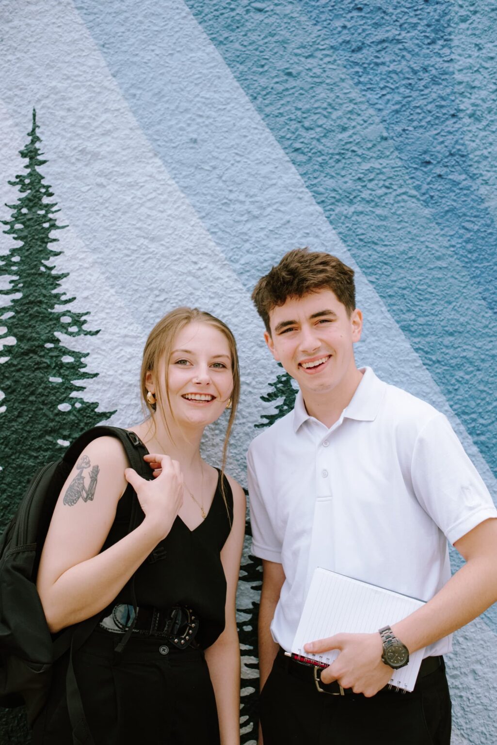 A young man and woman smiling in front of a blue mountain-themed mural.