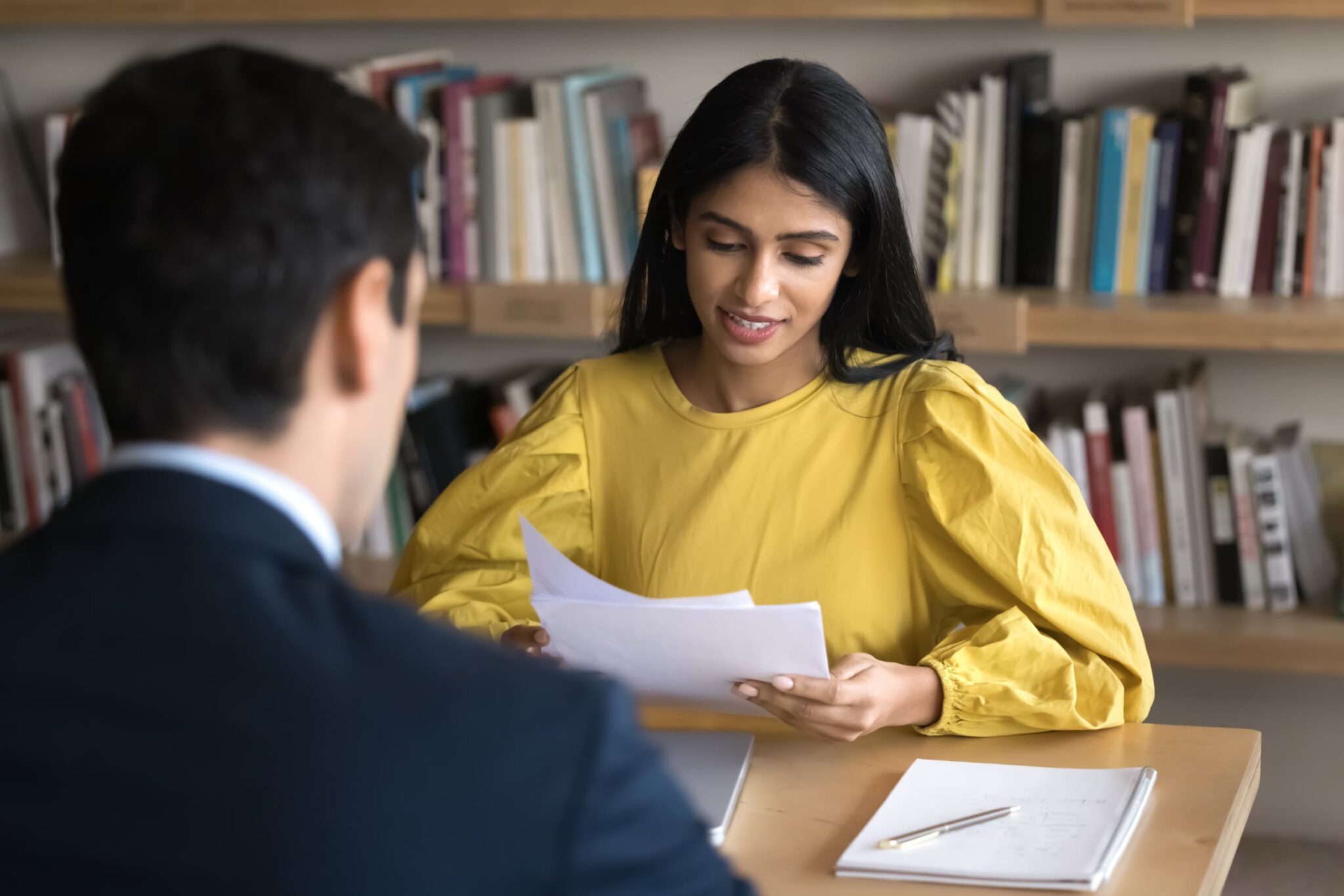 A woman in a yellow blouse reviews documents while sitting across from a person in a suit at a table in front of a bookshelf.