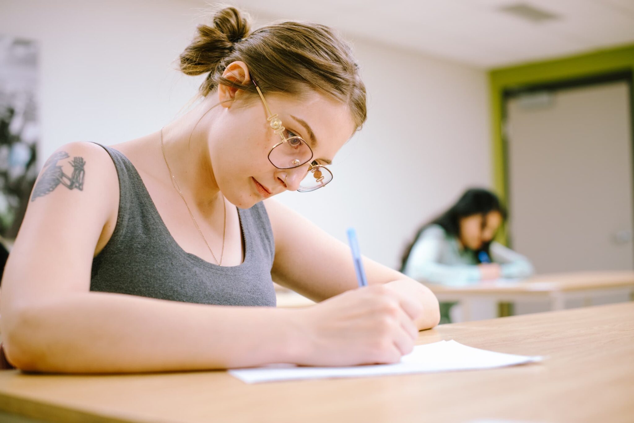 A woman with glasses writing on a paper at a desk in a classroom, with another student blurred in the background.