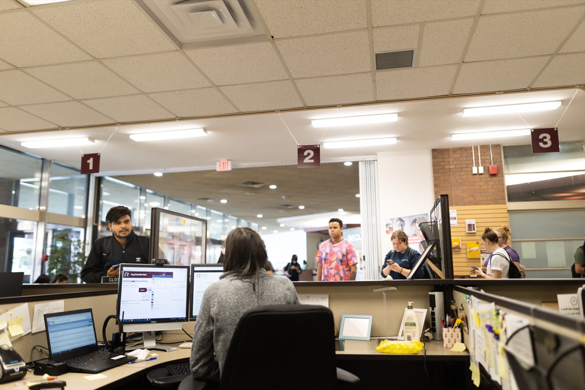 A service desk with staff at computers and students standing at a counter under numbered signs.