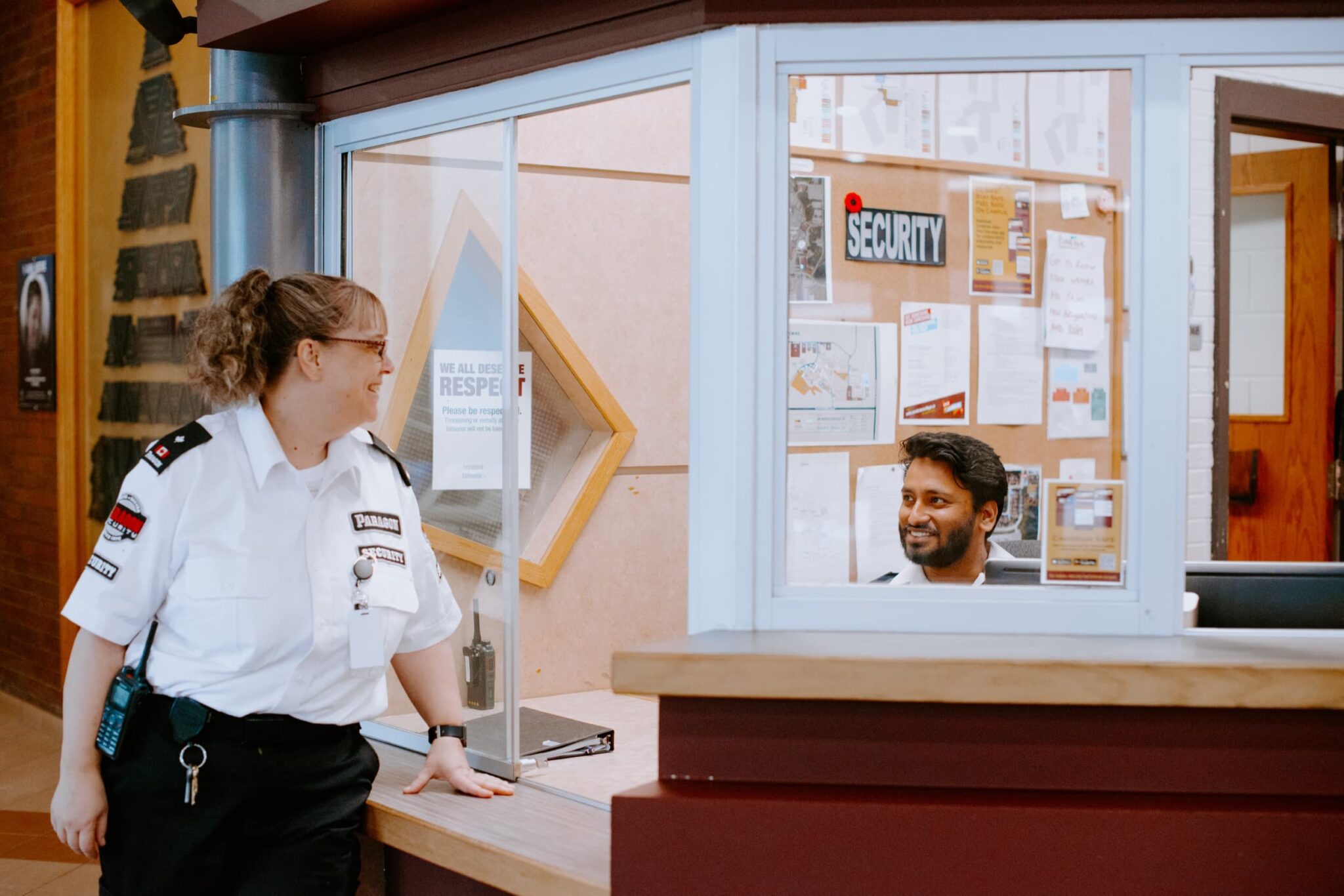 A security guard in uniform talking to a man sitting inside a glass-enclosed office.