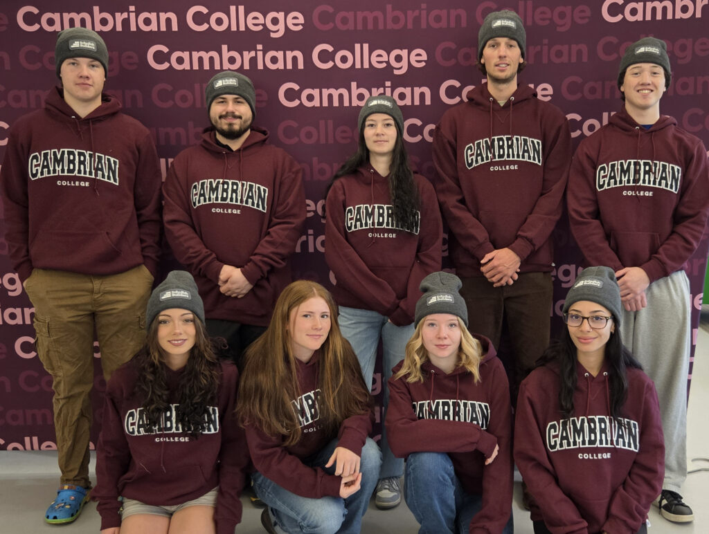 Nine students wearing matching maroon Cambrian College hoodies and grey beanies pose for a group photo in front of a branded backdrop.